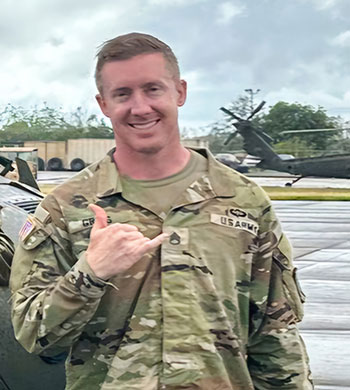 A smiling man in U.S. Army camouflage uniform standing outdoors on a military base