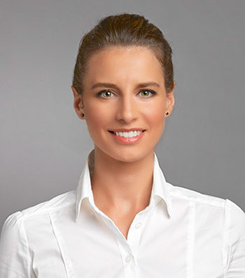 A professional woman smiling in front of a neutral gray background. She is wearing a crisp white button-down shirt with her brown hair neatly pulled back.